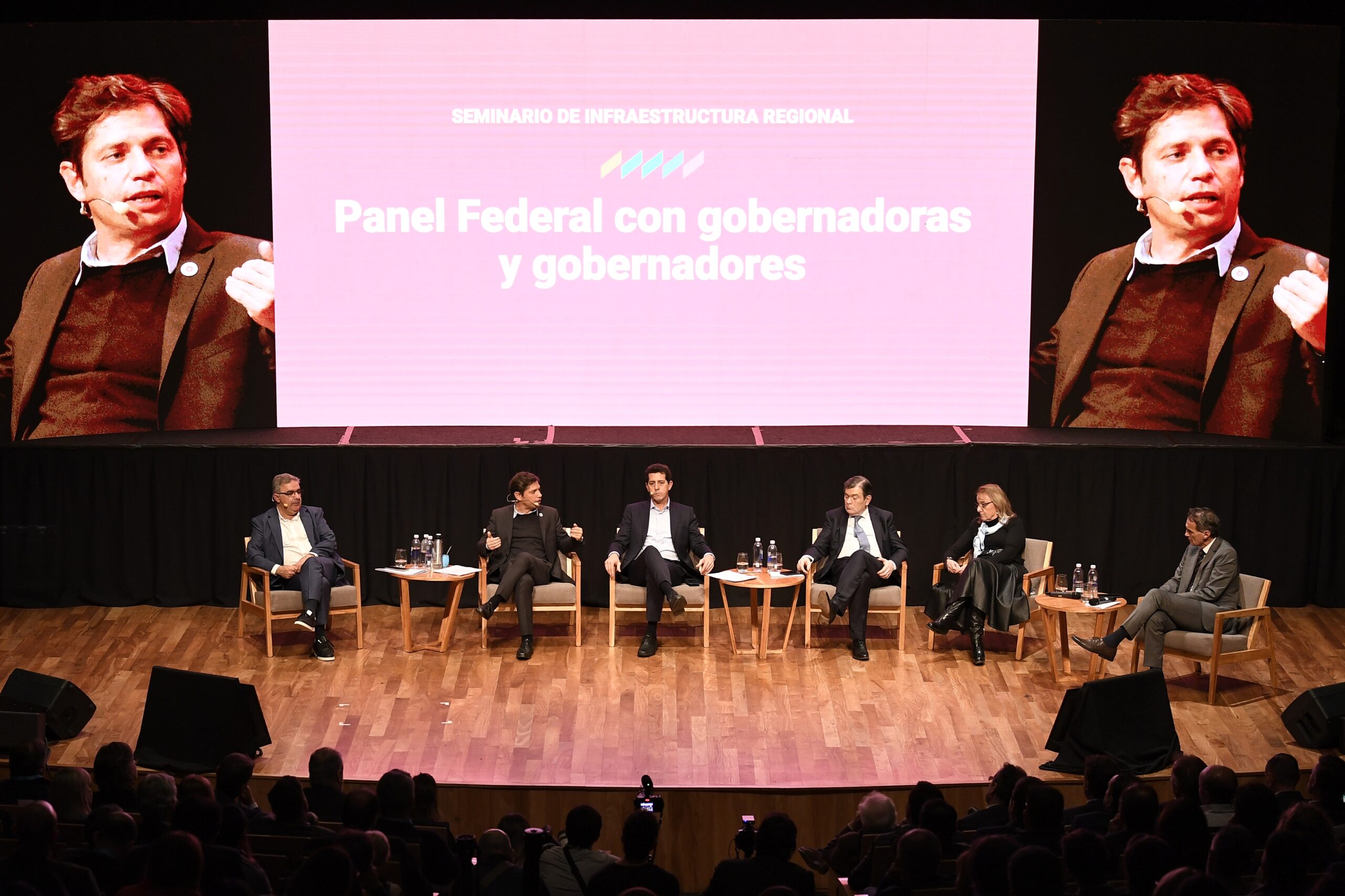 Kicillof hablando en el seminario sentado en una silla junto a otros oradores que lo escuchan